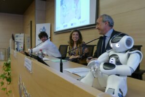 En la imagen, tres personas están sentadas en el salón de actos del Hospital San Juan de Dios de Córdoba durante la presentación de los Proyectos de Innovación Digital Next Generation aplicados a la Atención Temprana